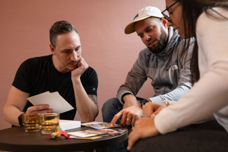 tre personer sitter vid ett bord och tittar på en yrkeskortlek.
