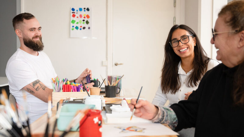 tre personer sitter vid ett bord och skrattar samt håller på och tecknar och målar.