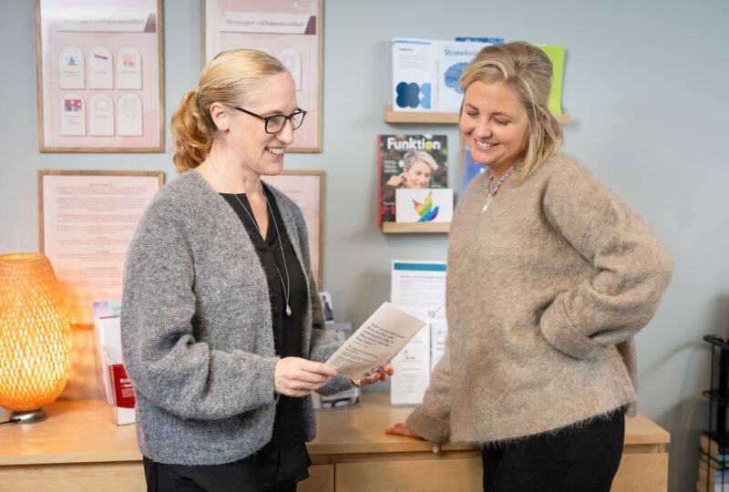 Två kvinnor med ett glatt leende står och tittar i broschyrer