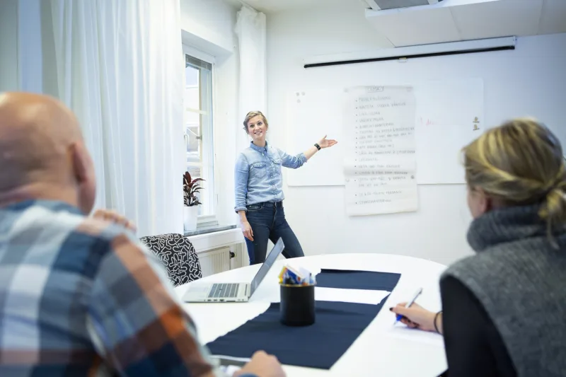 Fotoillustration: en Misa-anställd står vid en whiteboard på ett kontor och går igenom en punktlista för arbetsrutiner. Två arbetskonsulenter sitter vid ett konferensbord och lyssnar.