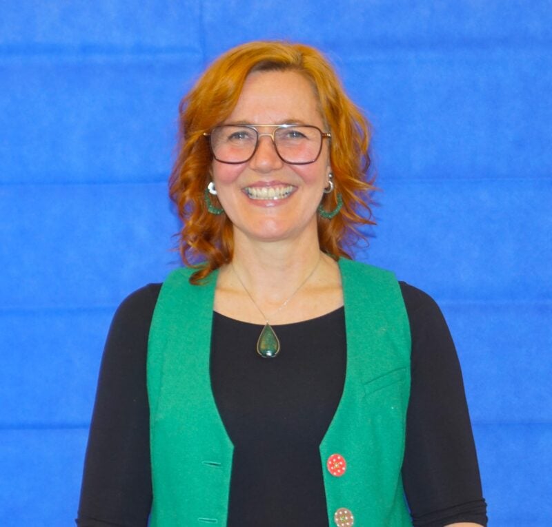 Kvinna med glatt leende och rött hår, står framför en blå vägg, hon har en svart tröja och en grön väst på sig-