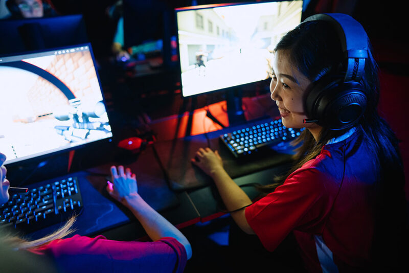 Två tjejer som spelar datorspel framför varsin skärm. Den ena pratar med den andra och ler.