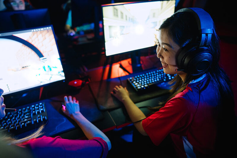 Två tjejer som spelar datorspel framför varsin skärm. Den ena pratar med den andra och ler.