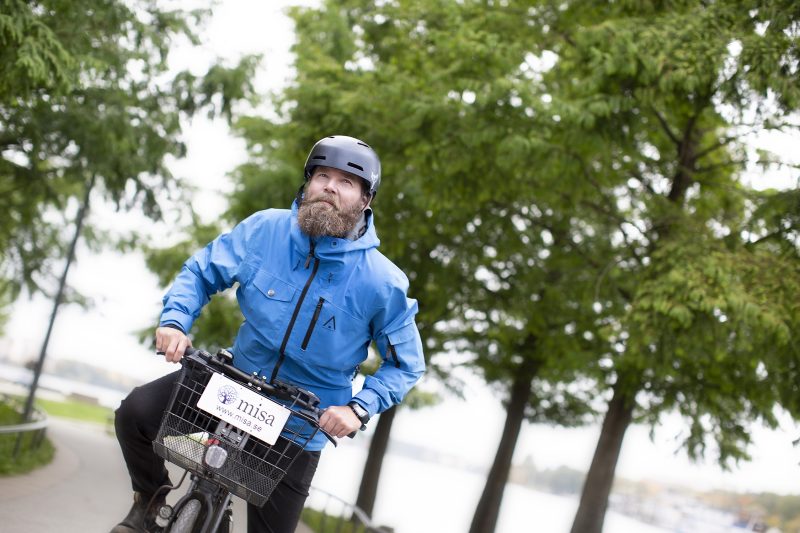 Man på cykel med hjälm en cykelkorg där det hänger en skylt med Misas logga