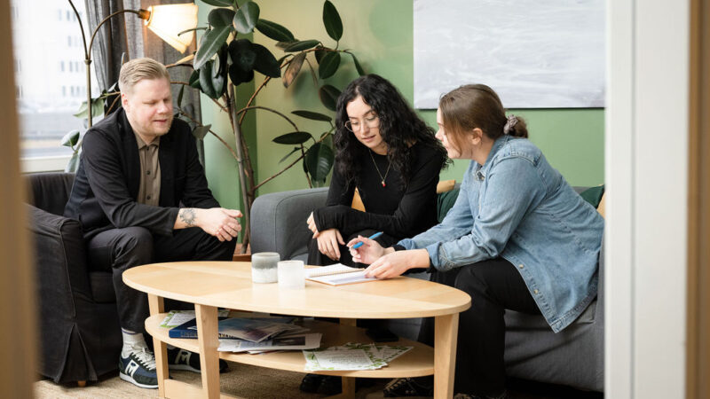 tre personer sitter runt ett lågt bord och det är en grön vägg och en grå soffa som de sitter i