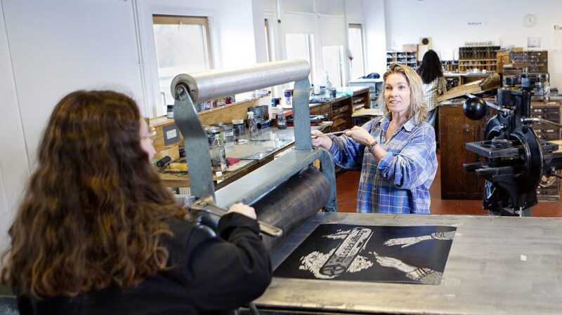 Två personer står vid en gammal hand tryckpress i Misa Konst och grafiks lokaler på Grafiksskolan