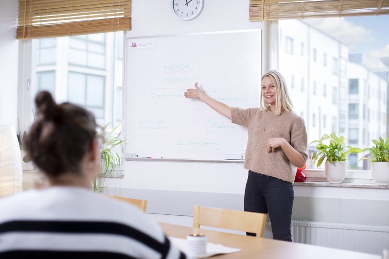 en kvinna står framför en whitebord och håller upp handen för att visa en text till den andra personen