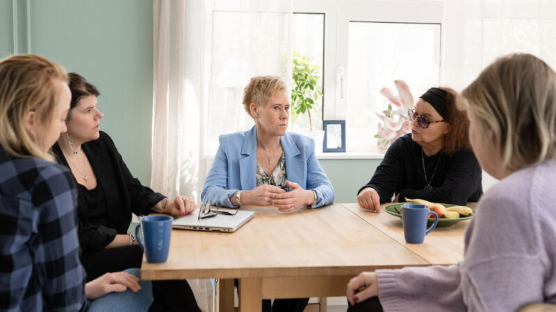Fem kvinnor sitter och pratar runt ett bord. bakom finns ett stort fönster.
