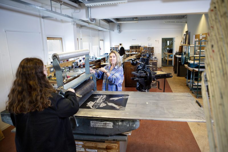 Två personer står vid en gammal hand tryckpress i Misa Konst och grafiks lokaler på Grafiksskolan