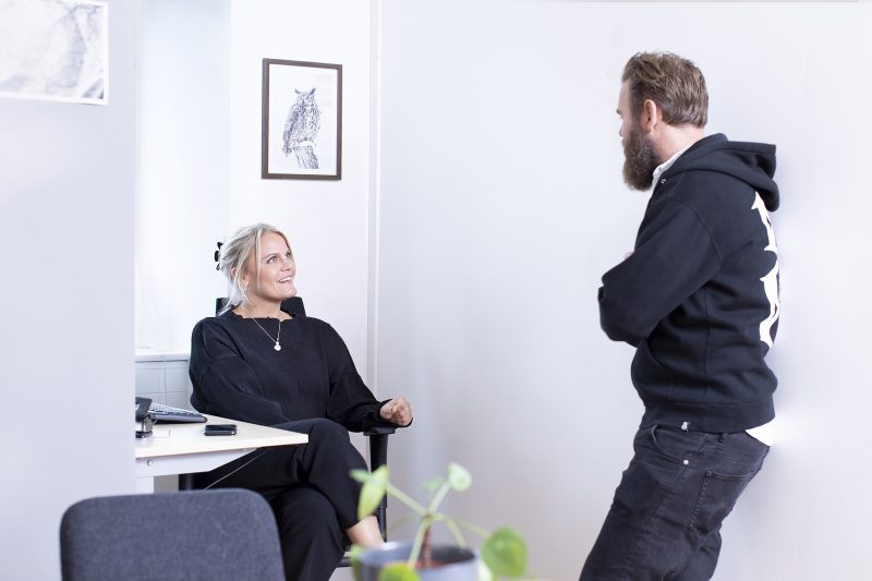 Två Misa medarbetare prtar en sitter vid ett skrivbord den andra står upp.