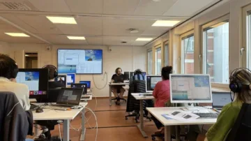Dataklassrum med flera datorer och en lärare med storskärm bakom sig.